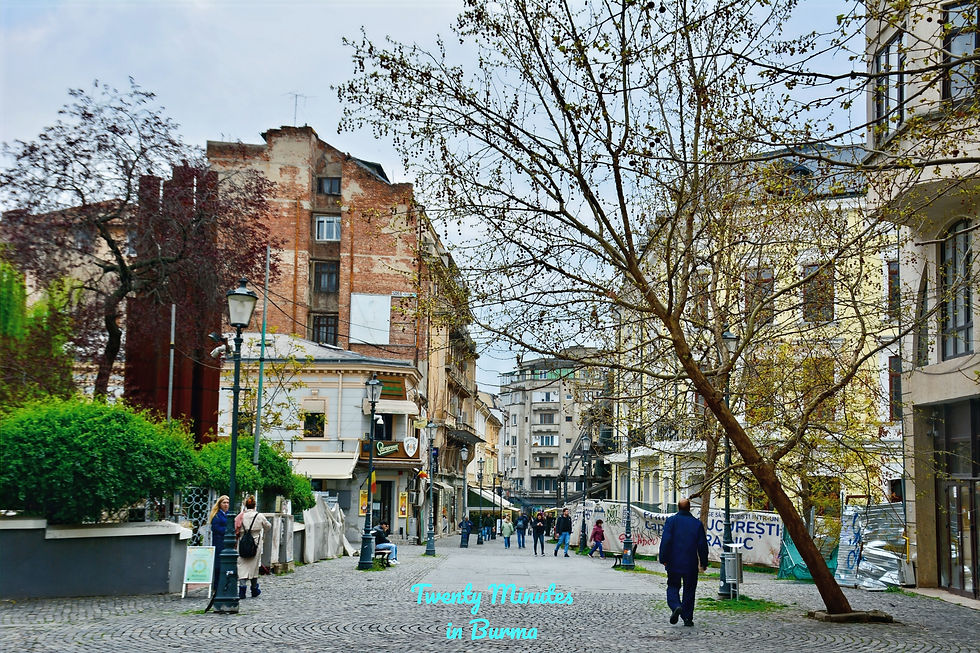 A cobblestone street with people walking, surrounded by historic buildings with red brick and yellow facades. Bare trees hint at early spring. Old Town, Bucharest, Romania