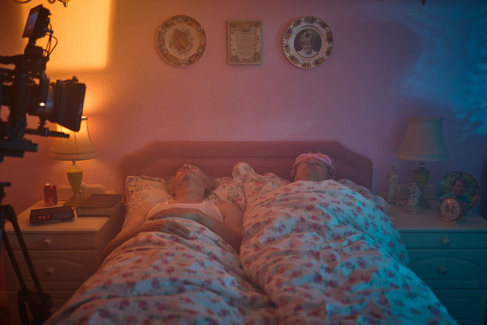 A couple sleep in a bed with a floral frilly bedspread, surrounded by decorative plates, the lords prayer and dated furniture.
