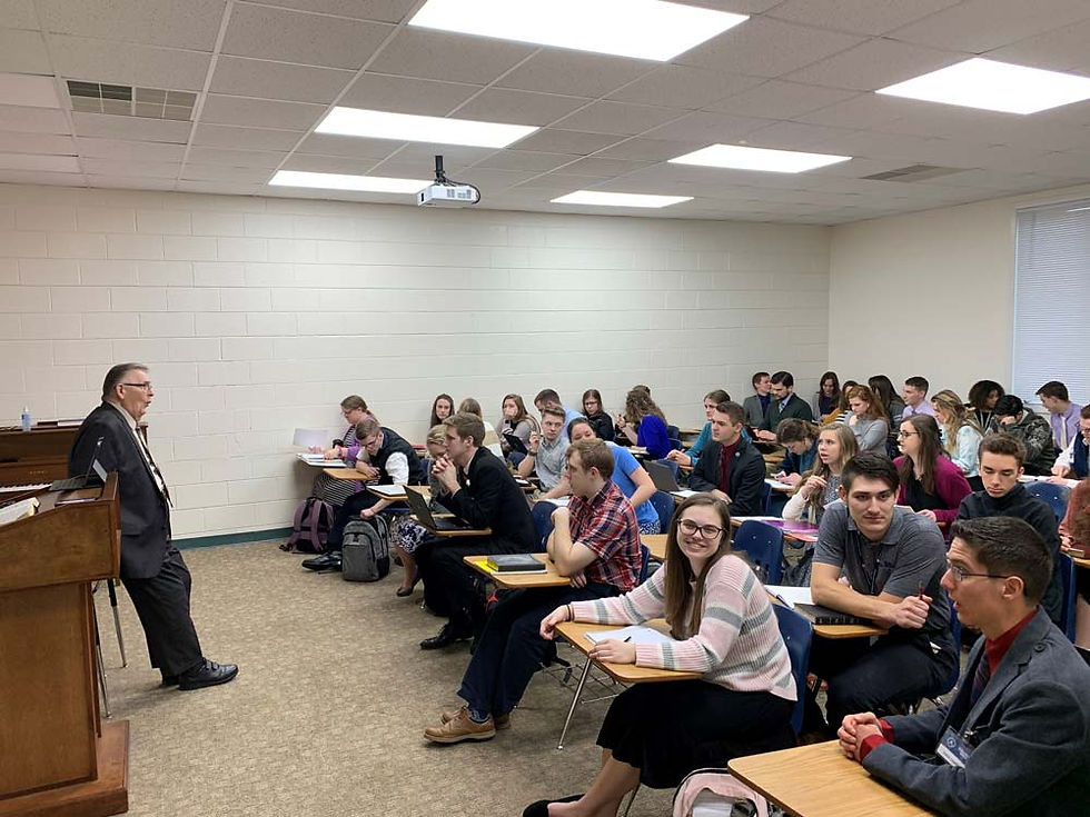 A teacher stands by a piano, lecturing to a full classroom of attentive students in casual attire. Bright, neutral-toned room.