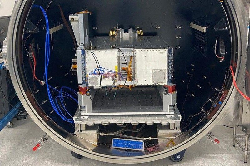 A white metal device with blue and orange wires is inside a large cylindrical chamber, against a gray industrial background.