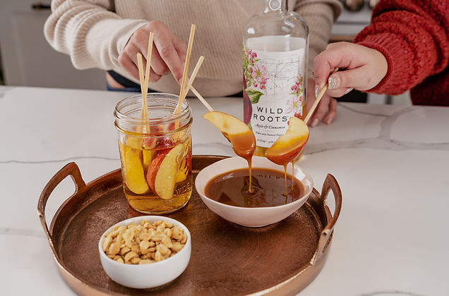 Apple slices being dipped in melted carmel on tray with nuts. Vodka in the background.