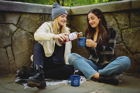 Two women sitting on blanket. One is holding Broken Top mug, while other is pouring Broken Top whiskey in the mug.