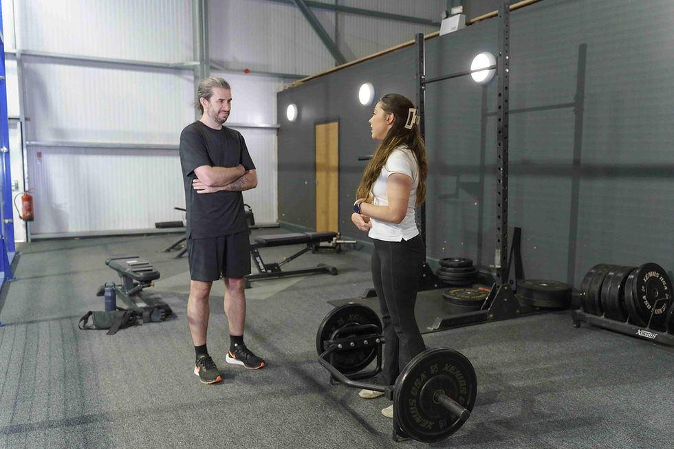Our osteopath and patient talking in a gym, with weights and equipment behind them. The osteopath stands near a barbell. The setting is bright and organised.
