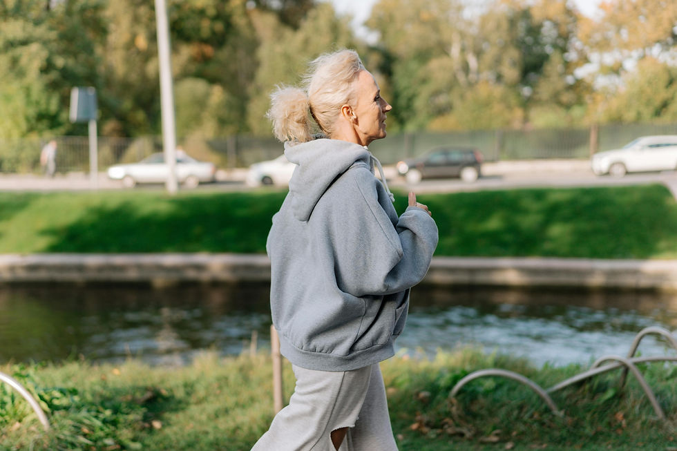 Woman jogging in a park, wearing a gray hoodie and sweatpants. Background shows lush greenery and a small river. Cars are on a distant road.