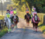 6 young people riding horses down a gravel road with a hedge on one side and a fence overlooking a field on the other. Two of the girls are wearing pink high visibility clothing and the others are wearing yellow