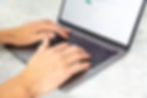 Hands typing on a gray laptop with a bright screen displaying blurred text. Set on a light marble-textured surface.