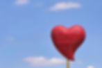 Red heart-shaped sculpture with triangle patterns on a pole against a bright blue sky with scattered clouds. Peaceful atmosphere.