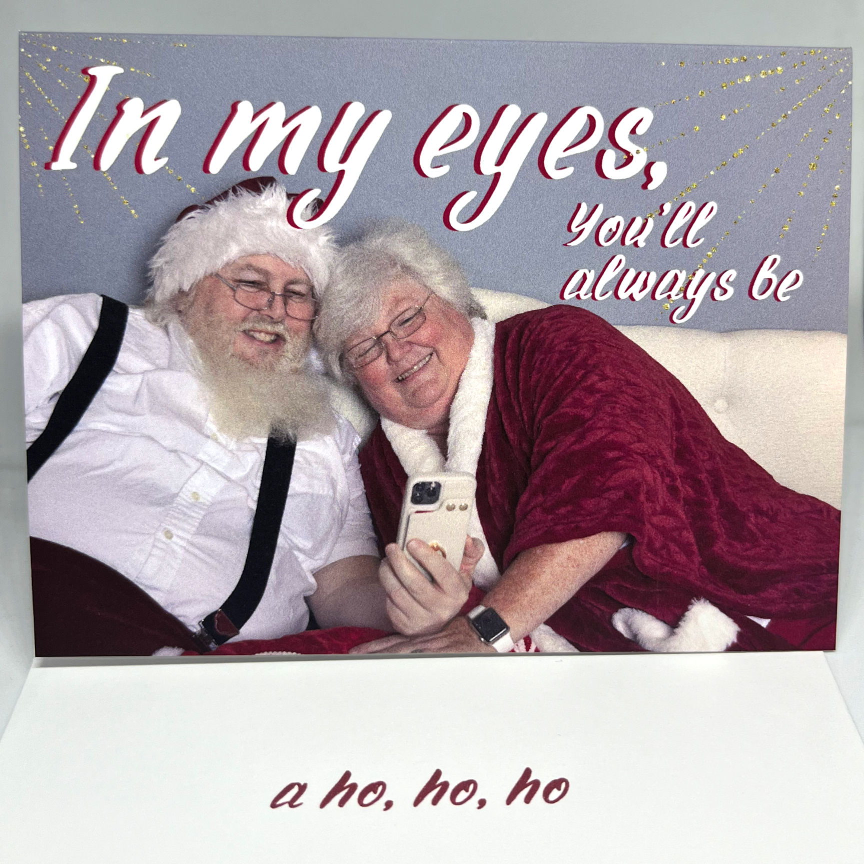 Greeting card featuring Santa and Mrs. Claus on a white sofa looking at a cellphone together smiling and cuddling with the text that says "In my eyes you'll always be" on the outside and "a ho, ho, ho" on the inside
