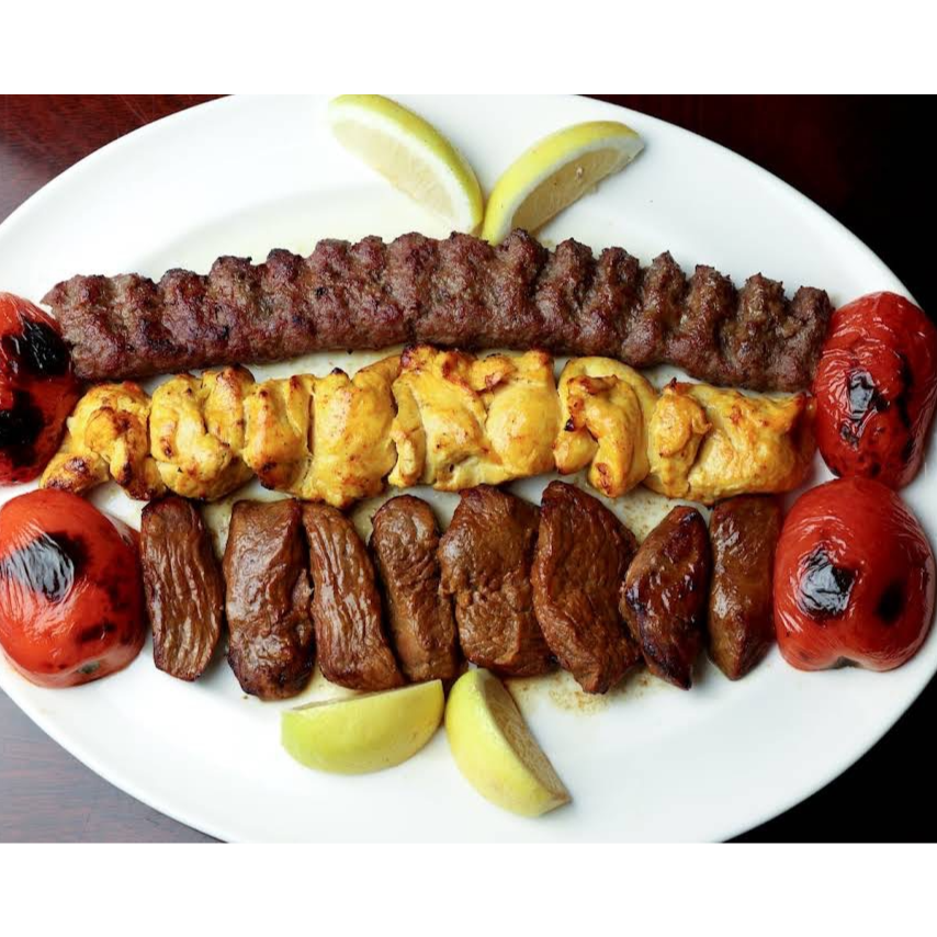 This image displays a hearty mixed grill platter featuring a long skewer of seasoned ground beef, marinated chicken cubes, and thick slices of grilled steak, all accompanied by two roasted tomatoes and lemon wedges. The arrangement suggests a generous and flavorful Middle Eastern meal ready to be served.