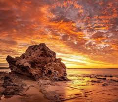 “Rock formation on sandy beach illuminated by dramatic red sunset sky.”