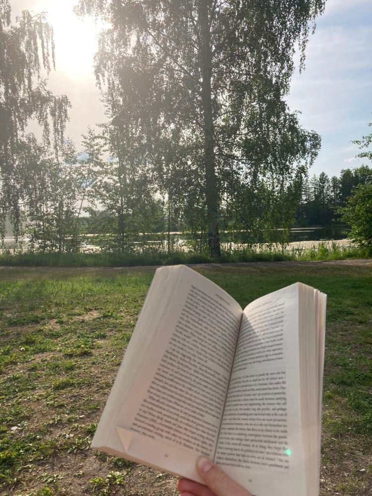 A book is held open by a hand in the foreground and the background shows a lake and tress in Tampere.