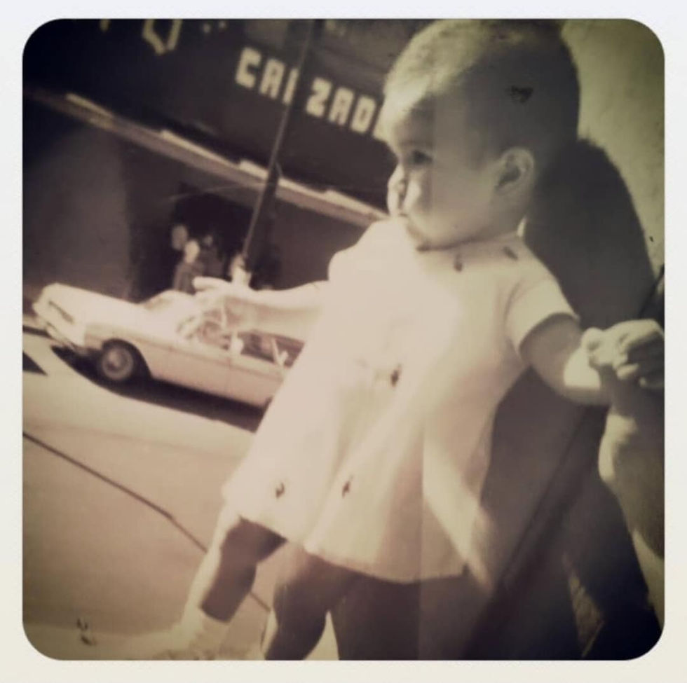 A sepia-toned photograph of a one-year-old girl standing on a balcony, wearing a white dress with small dark appliques. She’s reaching out with one hand while being gently supported by an unseen adult. A vintage car and a partial sign reading “CALZADO” appear in the blurred background, evoking an early nostalgic, 1970s-era setting.