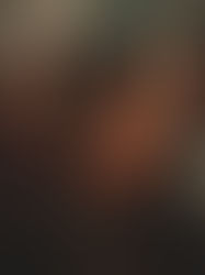 A woman with dark hair looks directly toward the camera while standing in a crowded space. Her face is softly lit, with blurred figures and warm tones surrounding her, creating a sense of presence and quiet intensity.