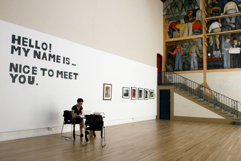 The artist seated alone in front of a gallery wall displaying the text ‘Hello! My name is …’ during the Hello! project, 2010, San Francisco.