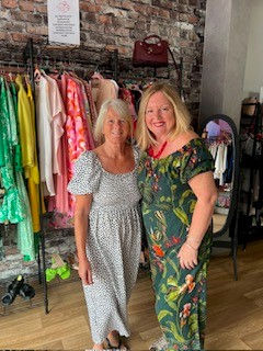 Two women standing and smiling in a boutique-style clothing shop, colorful dresses on racks, portrait orientation