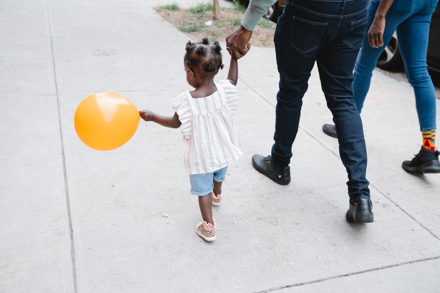 A toddler holding a balloon walks hand in hand with her parents on a city sidewalk