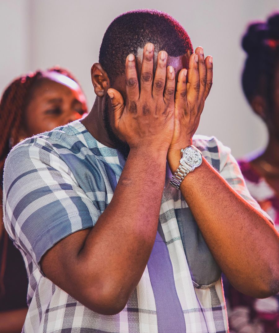 A Black man in church with his hands over his face in prayer, laying down a heavy burden