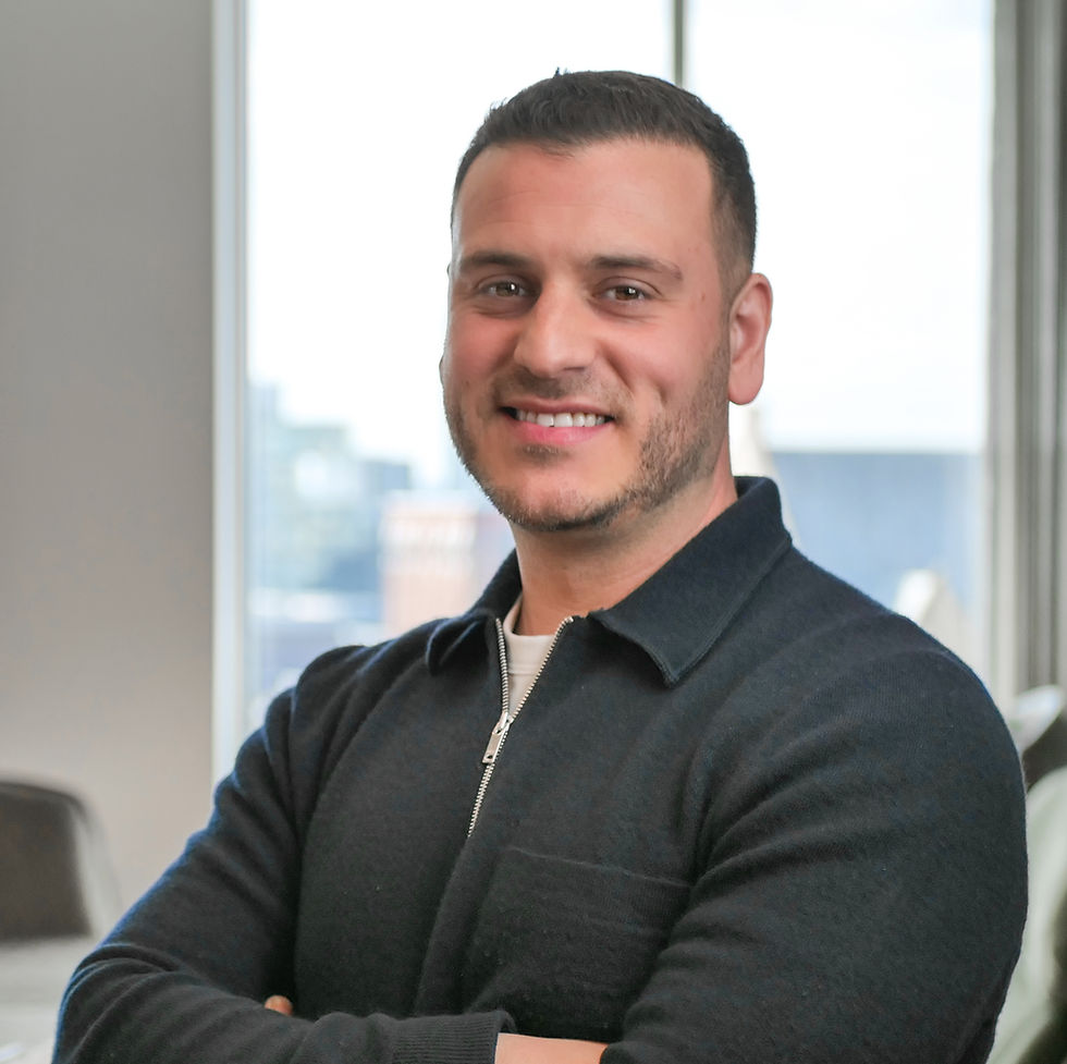 Jordan Saleh smiling with arms crossed, wearing a dark jacket in a modern office setting. Bright window in the background, conveying a positive mood.