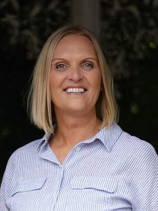 Tracey Field with short blonde hair wearing a light blue striped shirt. Dark, leafy background adds contrast to the cheerful expression.