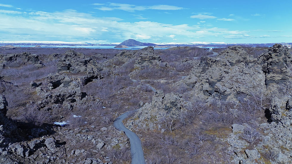 Rocky landscape with winding path, sparse vegetation, and a distant mountain under a blue sky. Snowy peaks visible on the horizon.
