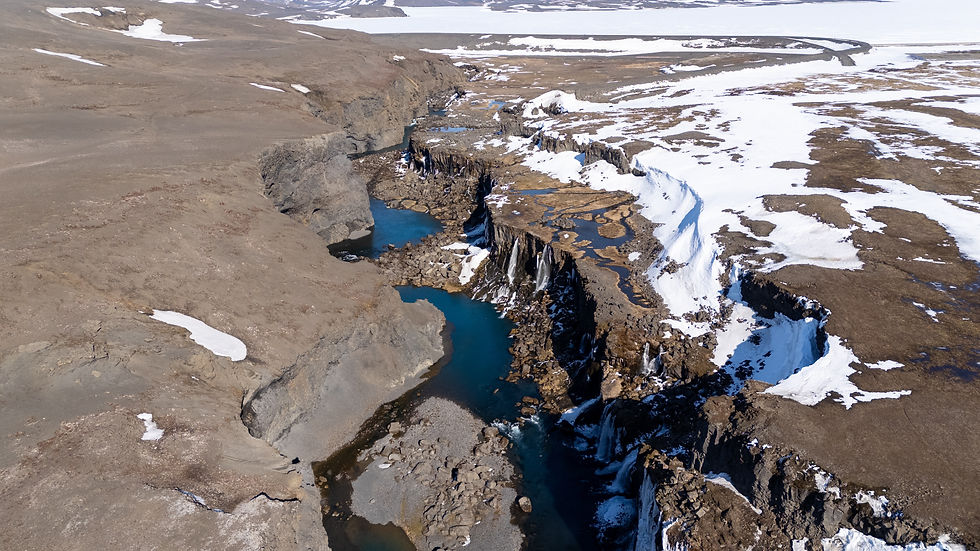 Aerial view of a canyon with a river flowing through rocky terrain. Snow patches scatter across the landscape, contrasting with blue water.