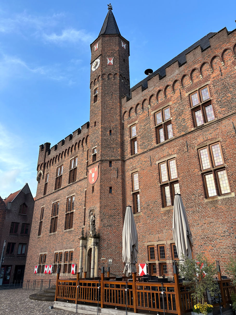 Historic brick castle-like building with a clock tower under a bright blue sky, with closed café umbrellas in front.