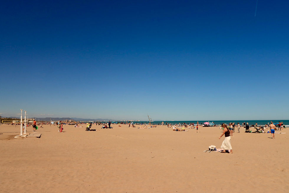 Crowded beach under clear blue sky, people sunbathing and strolling, colorful umbrellas, distant mountains, relaxed and sunny atmosphere.