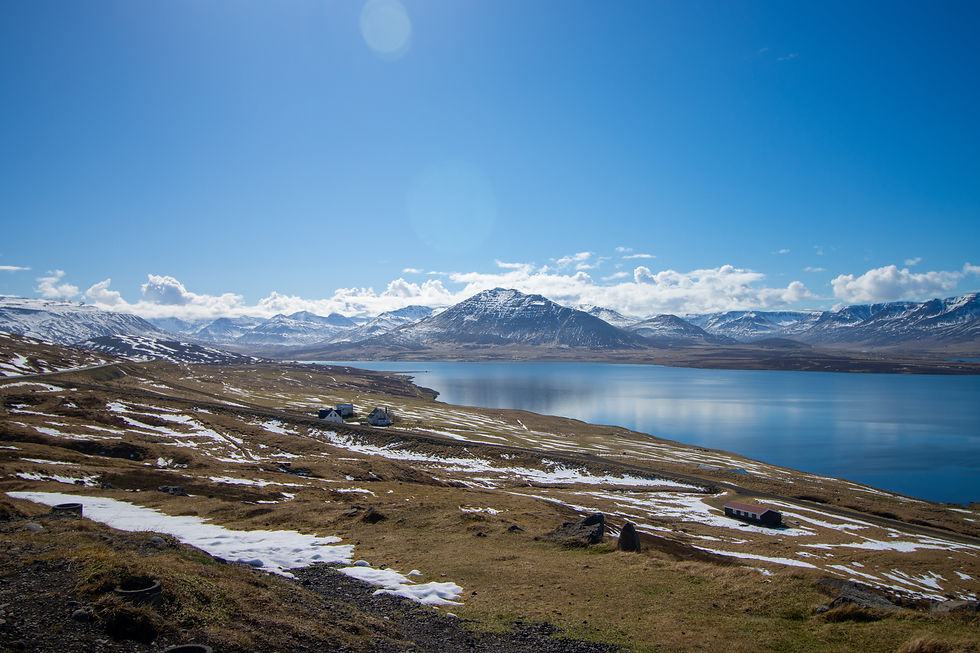 Snow-dusted mountains beside a calm lake under a bright blue sky. Sparse houses dot the grassy, sunlit landscape. Peaceful and serene.