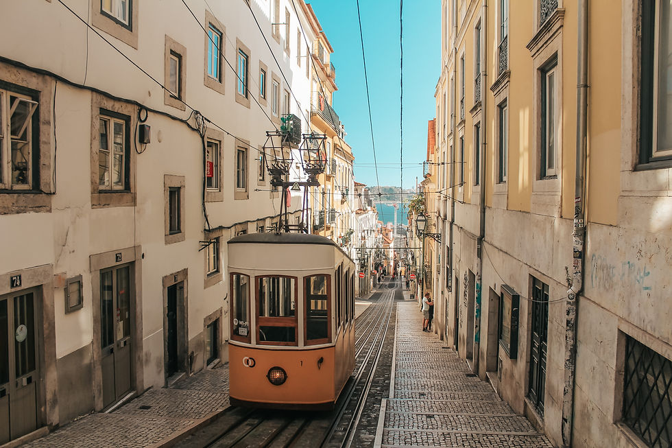 Historic tram on cobbled street in sunny city. Tall buildings line the street; distant blue water visible. Peaceful urban scene.