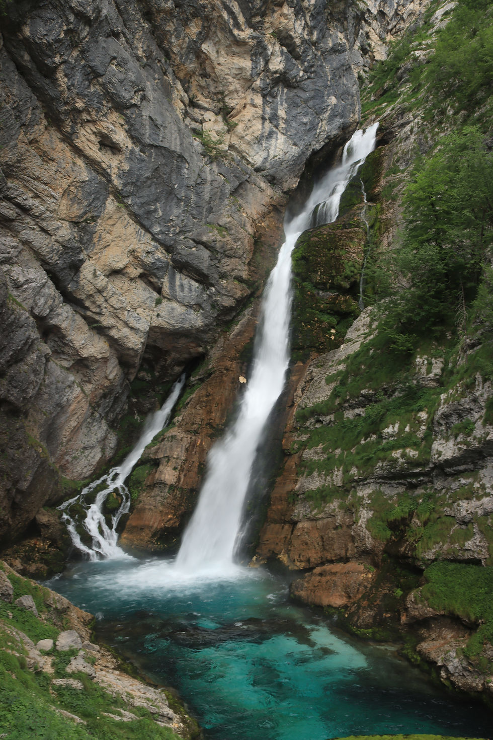 Tall waterfall pours through rocky gorge into turquoise pool, with mossy cliffs and green trees surrounding it.