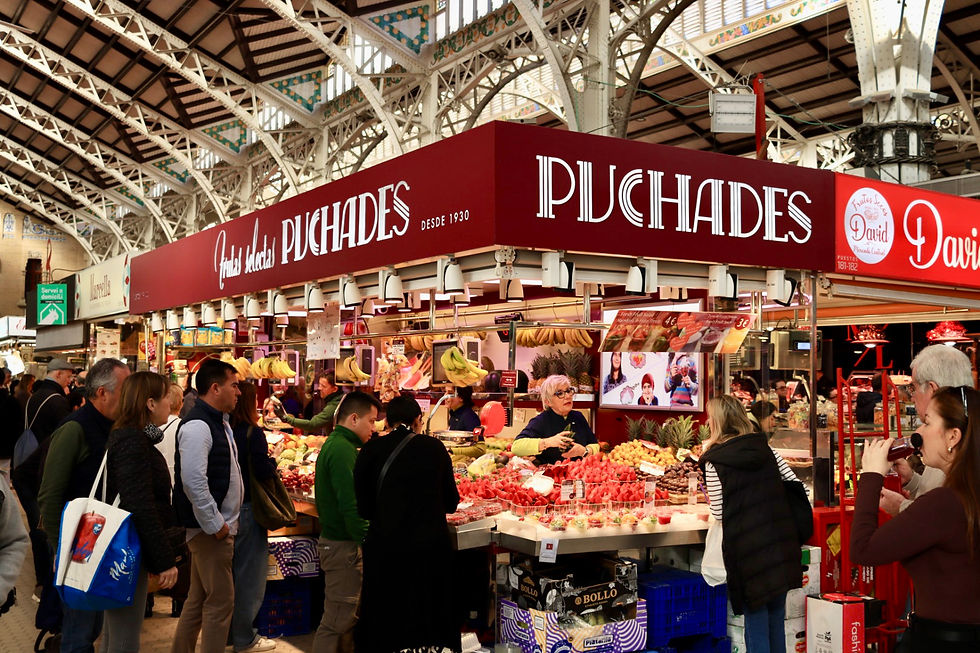 Crowd at market stalls labeled "Puchades," with various fruits and vendors interacting. The setting is vibrant, under an ornate roof.