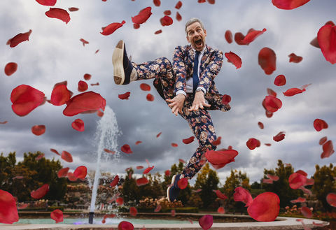 A joyful groom in a bold floral suit leaps high in the air during a Castle Farms wedding, surrounded by a swirl of red rose petals. A fountain sprays behind him under a cloudy sky, adding drama to the playful, mid-air moment.