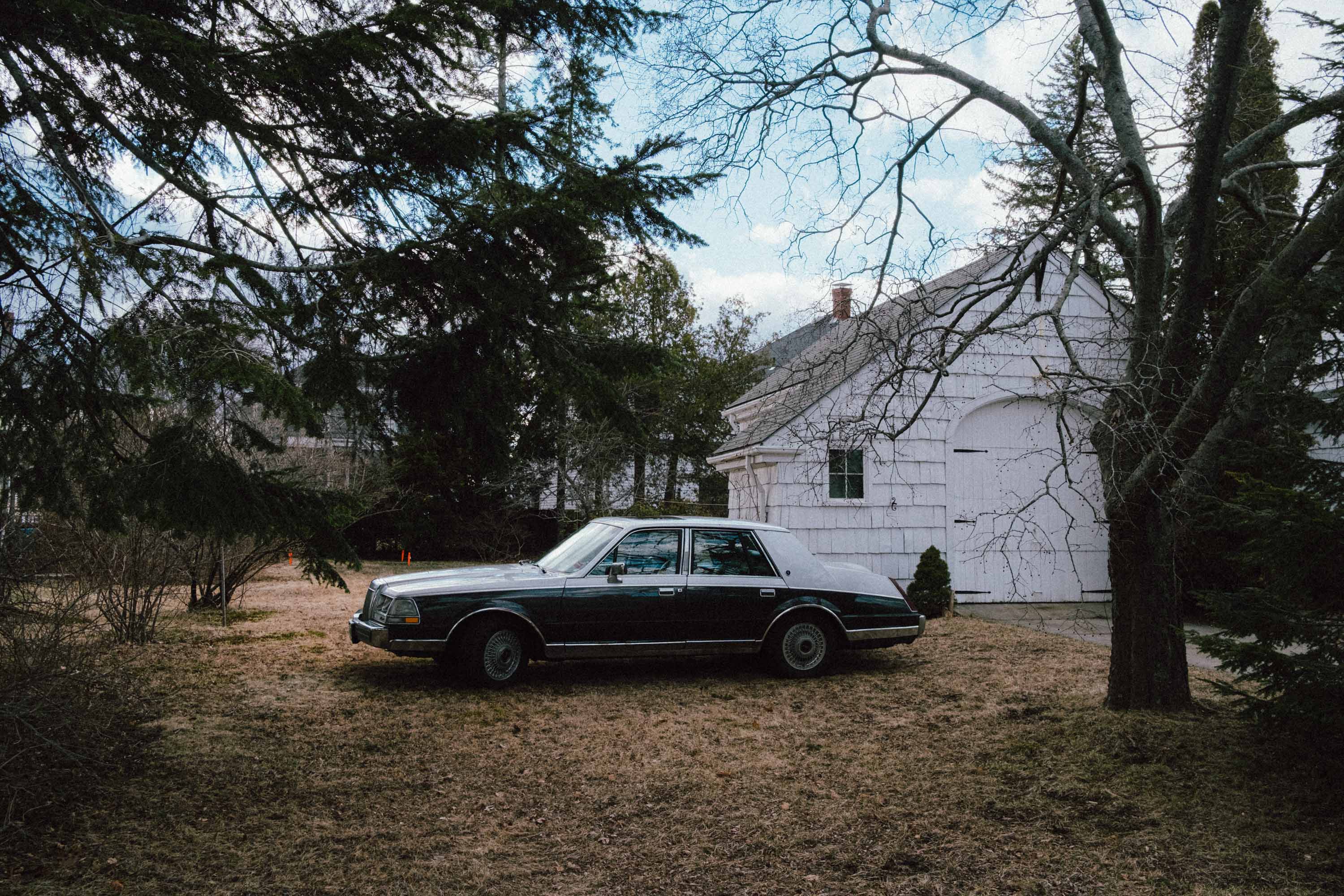 Voiture classique garée devant une maison en bois blanc et des arbres.
