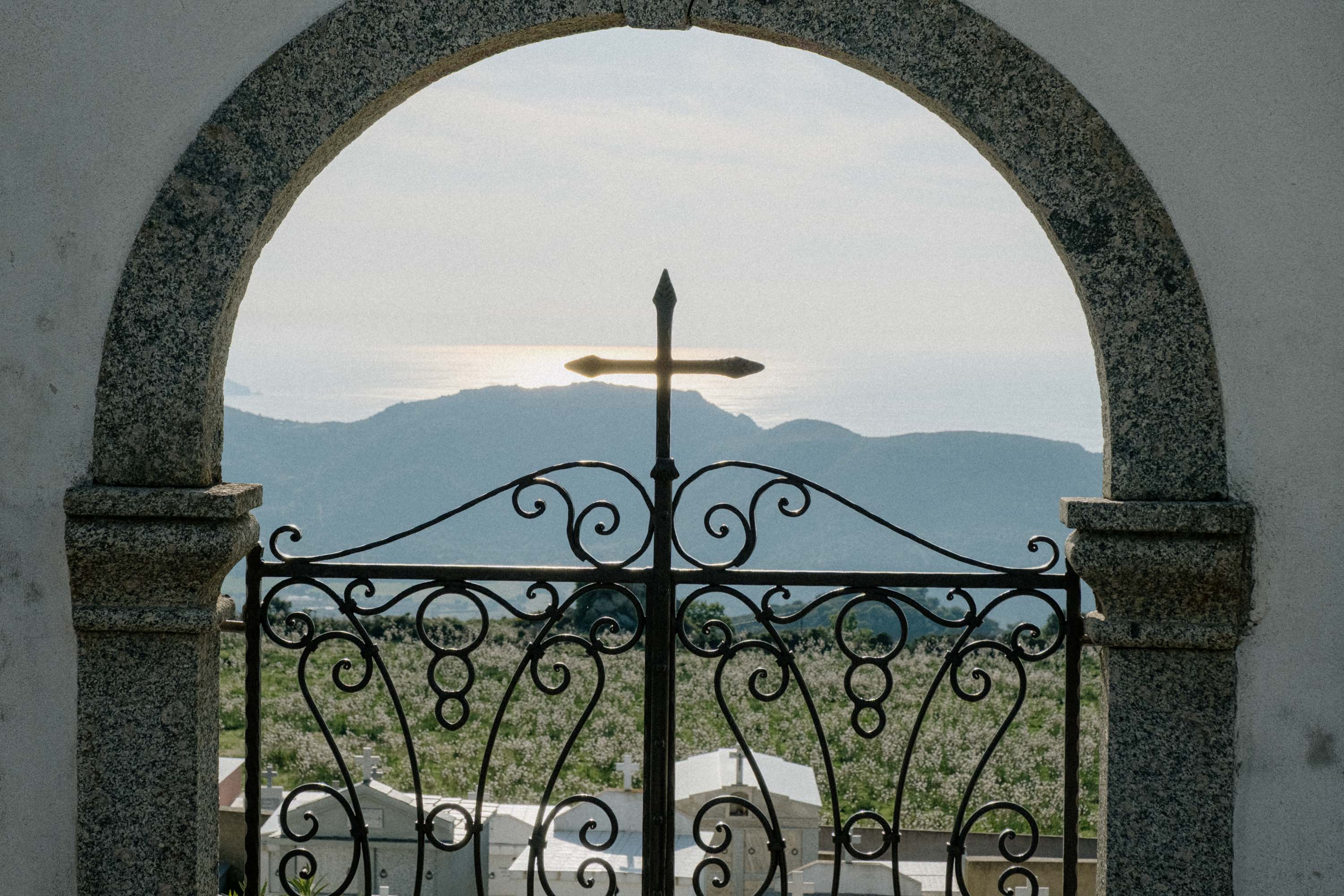 Croix sur portail en fer forgé devant un paysage montagneux et la mer.