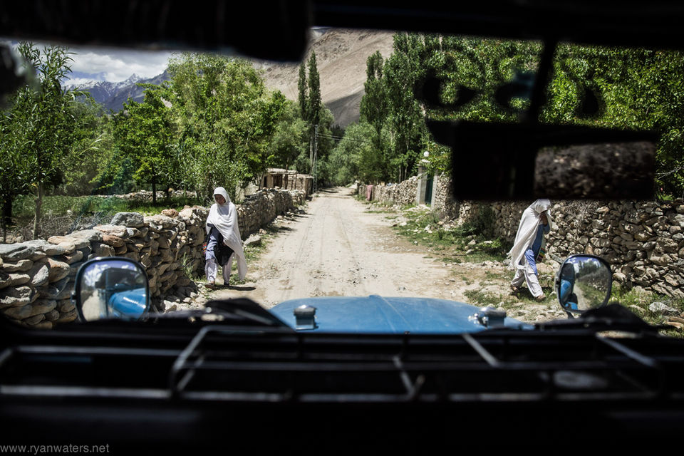 View from inside a vehicle driving down a rural dirt road lined with stone walls and trees, with two pedestrians walking along the roadside in a mountain village.