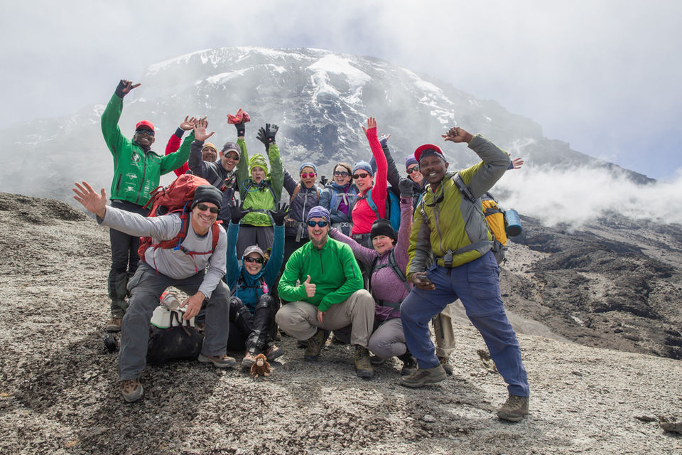 Group of about 8-10 trekkers celebrate at what appears to be a summit or high-altitude viewpoint, with arms raised in celebration, wearing colorful rain gear and outdoor clothing with misty mountains visible in the background.