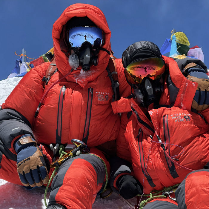 Two mountaineers in bright red expedition suits and oxygen masks pose for a selfie at high altitude. Snow-covered mountain peaks are visible in the clear blue sky behind them.