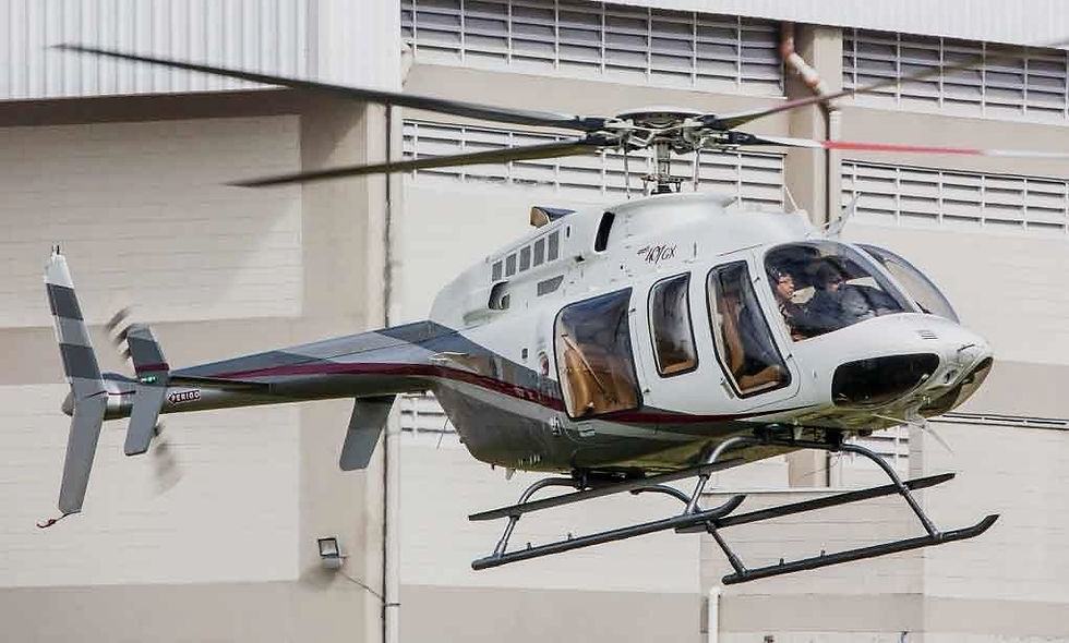 A white and grey Bell 407GX helicopter with a red stripe hovers in front of a light-colored building.