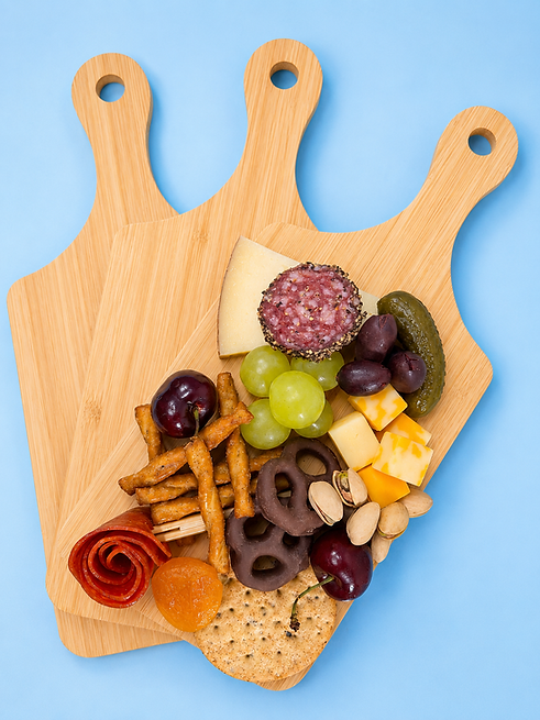 Three stacked and fanned mini charcuterie wooden boards with handle, the top one has charcuterie ingredients like cheese, cured meats, buts, pretzels and fruit.