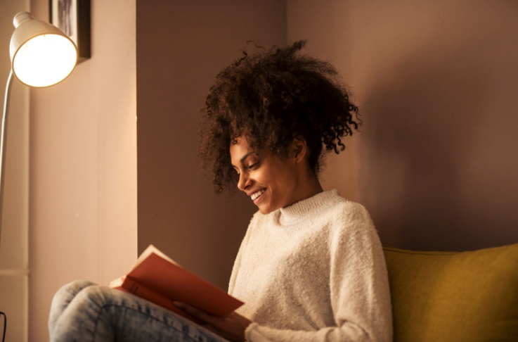 Black woman reading a book on a green sofa under a lamp.