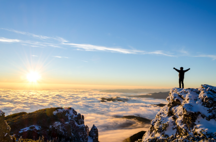 Man stands on mountain top with hands raised looking towards the sun.