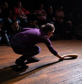 A performer in a purple shirt crouches low on a wooden stage, reaching toward a prosthetic arm lying on the floor. Audience members sit close around the performance area in the dimly lit theater, watching the intimate moment.