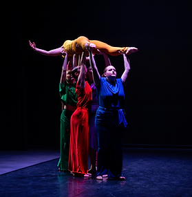 A group of four dancers in colorful costumes perform a lift on a dark stage, with three dancers in green, red, and blue supporting a fourth dancer in yellow who is held horizontally overhead. Dramatic purple and blue stage lighting highlights the dancers against a black background.