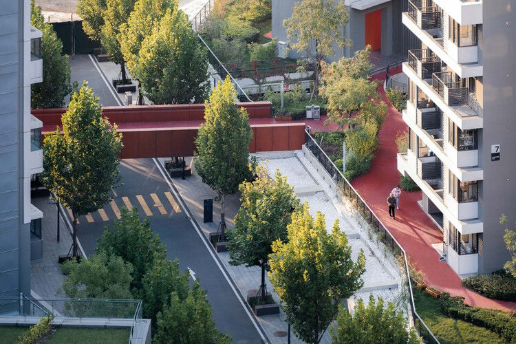 Aerial view of a modern urban park with trees, red walking path, crosswalk, and a person walking. Buildings surround the green space.