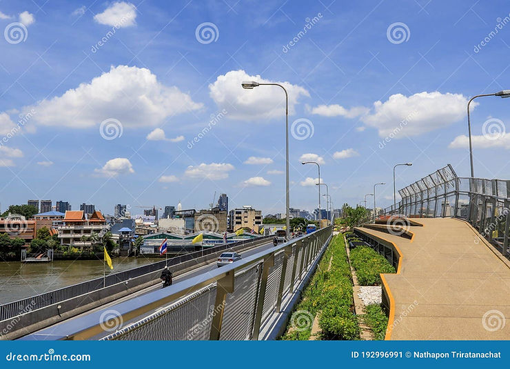 Pathway on a bridge with street lamps, overlooking a river and city skyline. Blue sky with clouds, Thai flags visible. Peaceful atmosphere.