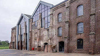 Historic brick building with large glass windows, featuring arched panes. Overcast sky, greenery in the background, and a red object.