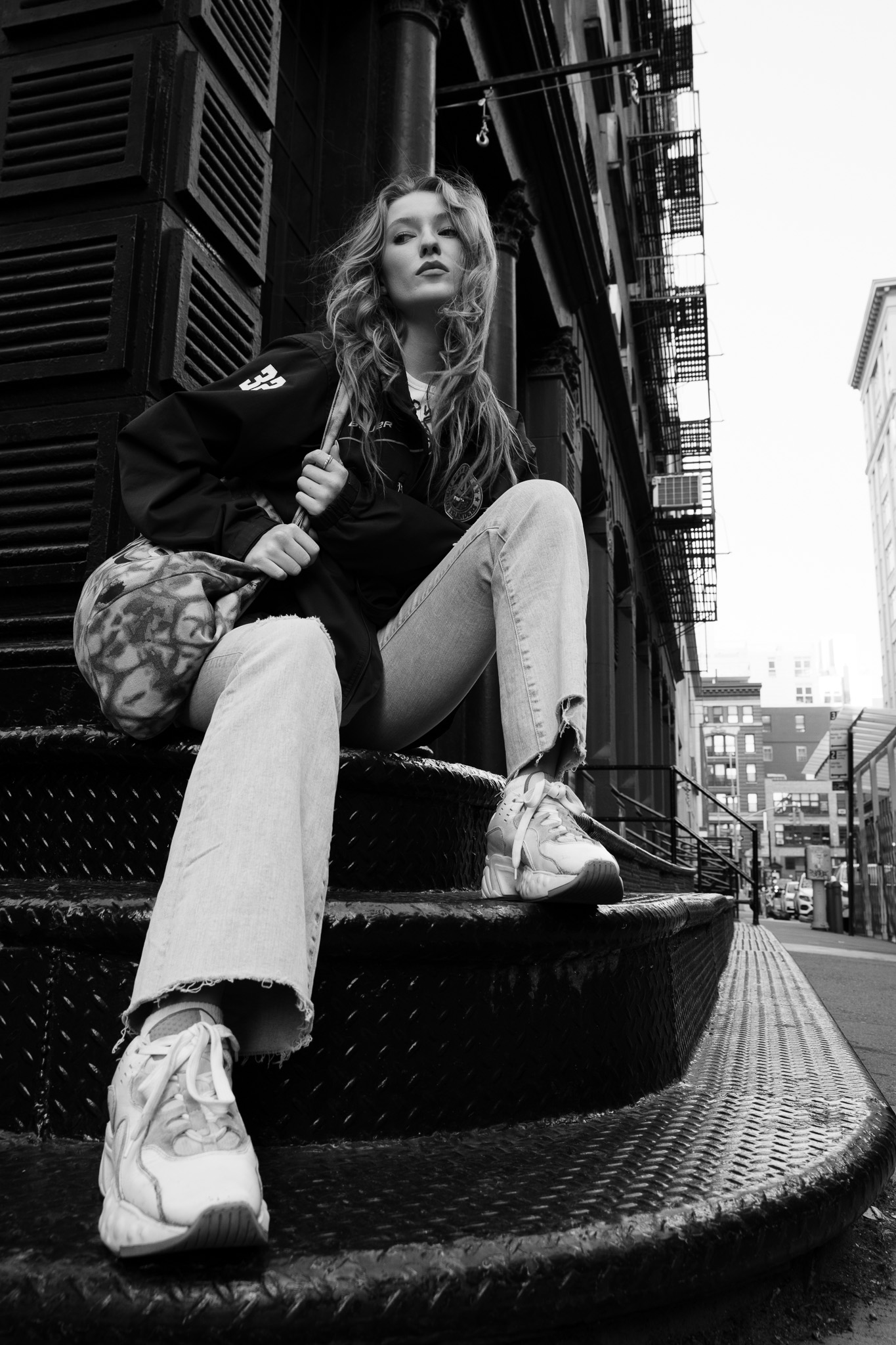 Black-and-white editorial portrait of a woman seated on city steps beneath fire escapes.