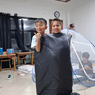 Two children stand together indoors inside a large black sack during a game, smiling at the camera. Another child sits inside a small play tent in the background.
