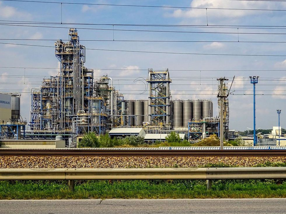 Industrial chemical plant with tall pipes and silos behind train tracks; Borealis sign visible under a bright blue sky.