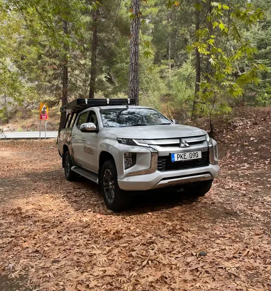 Front side of Mitsubishi L200 Tarasso Camper Truck, with closed rooftop tent in forest full of leaves on the ground